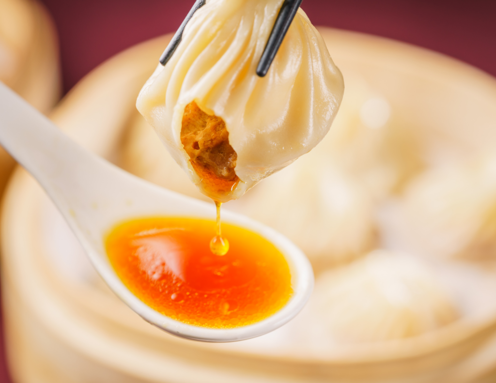 A photo of a soup dumpling being held with chop sticks with a spoon underneath catching the soup.