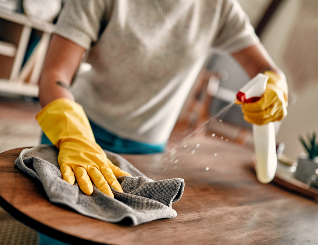 A person wearing yellow cleaning gloves, uses a cloth and cleaning solution to wipe down a table.