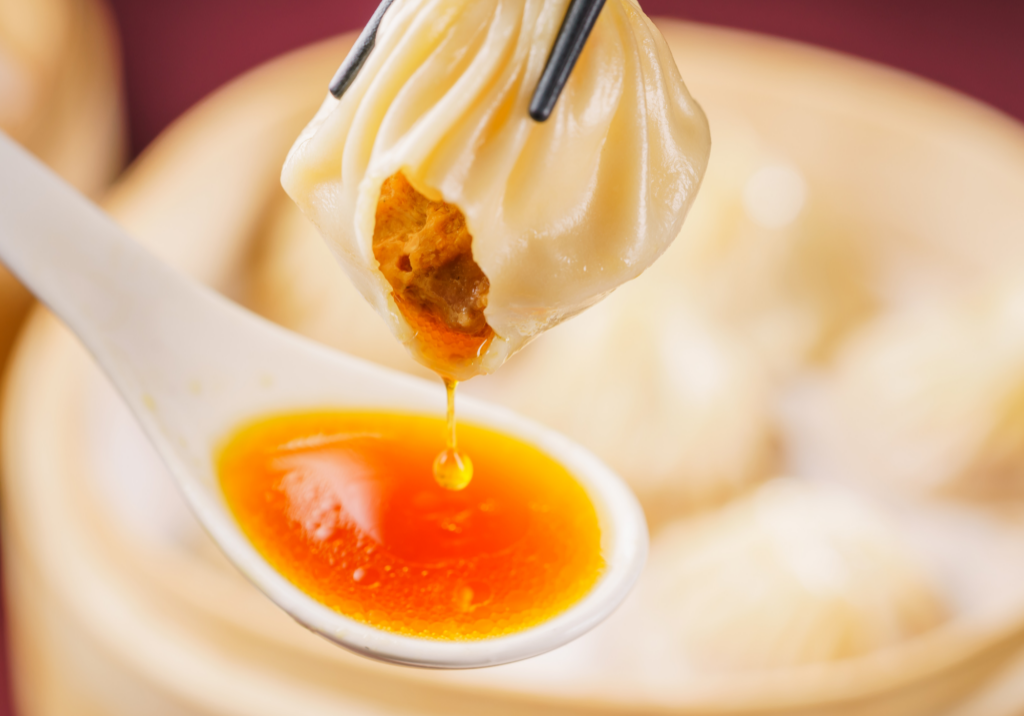A photo of a soup dumpling being held with chop sticks with a spoon underneath catching the soup.